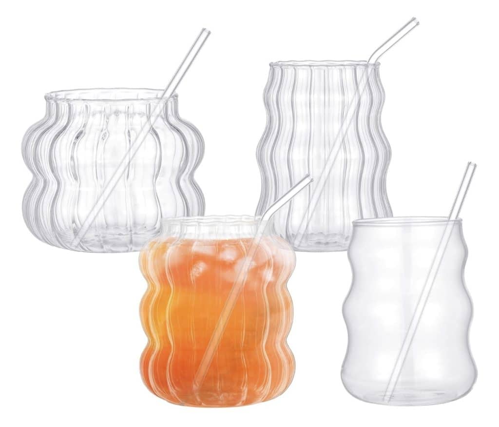 Four uniquely-shaped clear glass cups with transparent glass straws, three empty and one filled with an orange beverage and ice, are arranged against a white background—perfect for serving my favorite summer drinks.